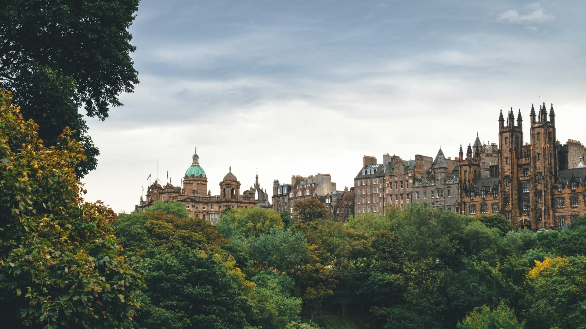 Edinburgh skyline rising above lush green parkland, with historic stone buildings and a domed roof under a soft summer sky